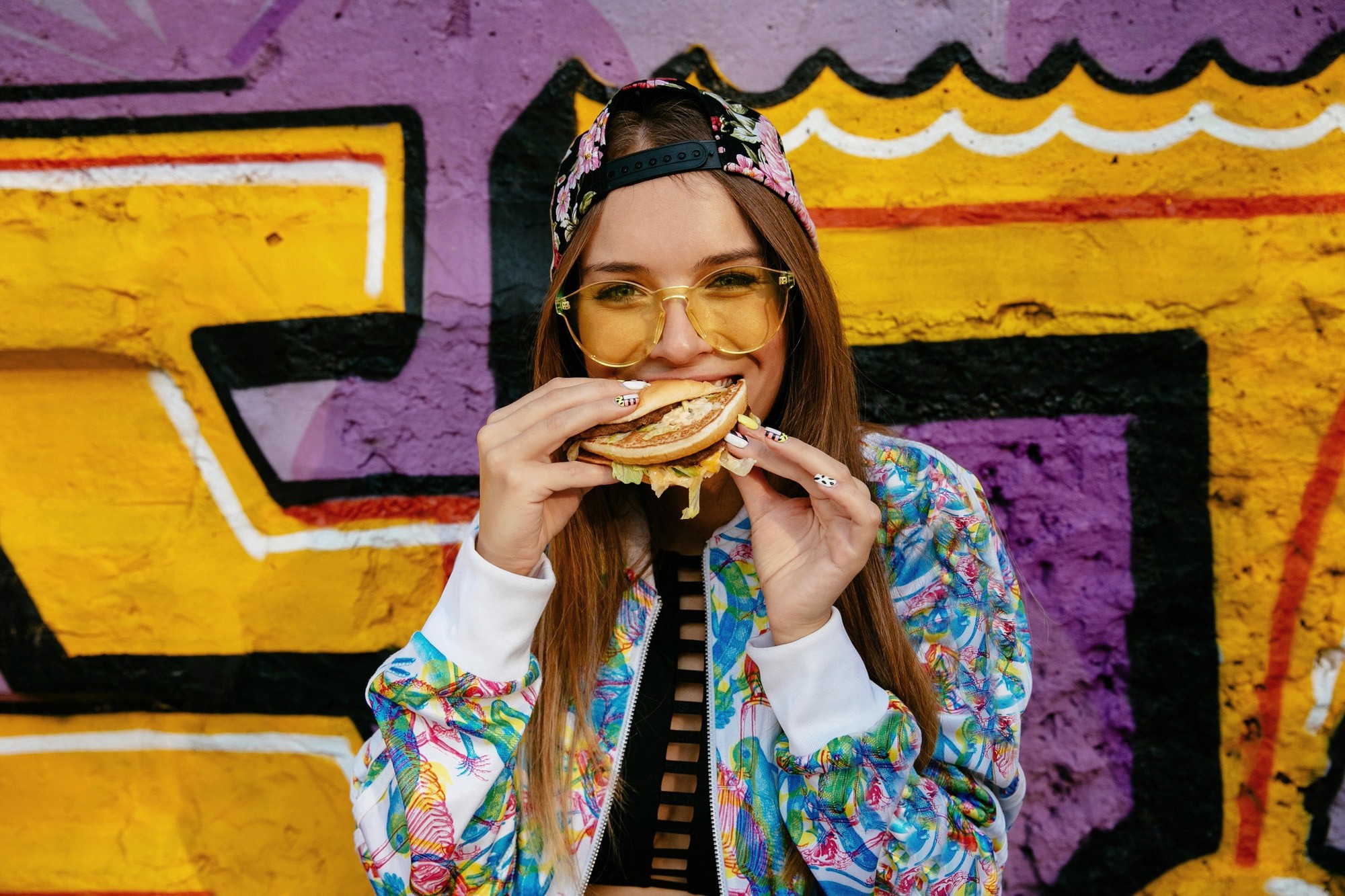 A young woman wearing yellow sunglasses and a colorful jacket taking a bite out of a burger in front of a graffiti wall.